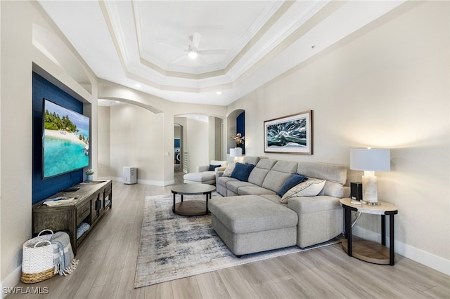 living room featuring arched walkways, crown molding, a raised ceiling, light wood-style floors, and baseboards