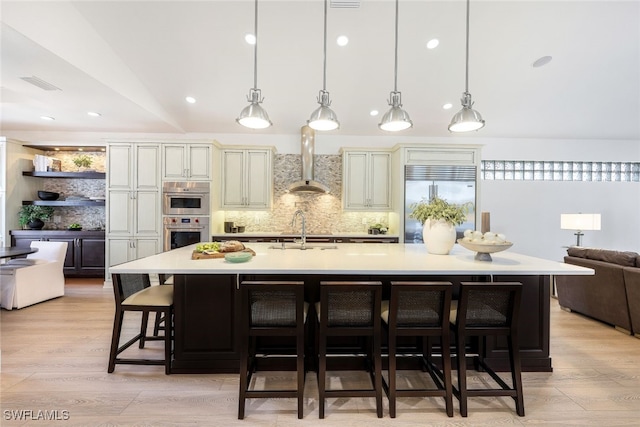 kitchen with hanging light fixtures, stainless steel appliances, wall chimney exhaust hood, cream cabinetry, and a large island with sink