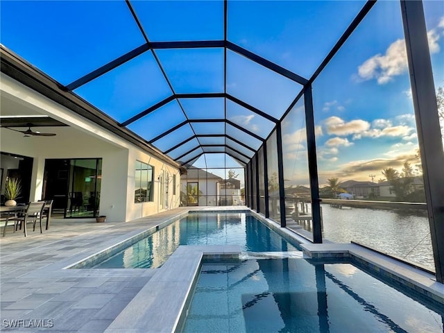 pool at dusk featuring an in ground hot tub, ceiling fan, glass enclosure, a water view, and a patio area