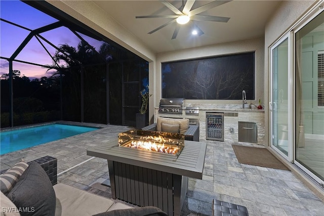 patio terrace at dusk with ceiling fan, an outdoor fire pit, wine cooler, grilling area, and an outdoor kitchen