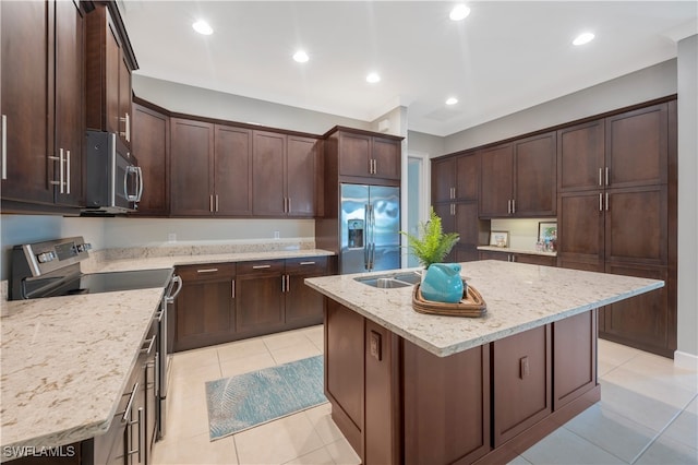 kitchen with light tile patterned flooring, appliances with stainless steel finishes, an island with sink, dark brown cabinetry, and light stone counters