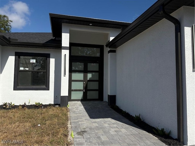 entrance to property featuring decorative driveway, roof with shingles, and stucco siding