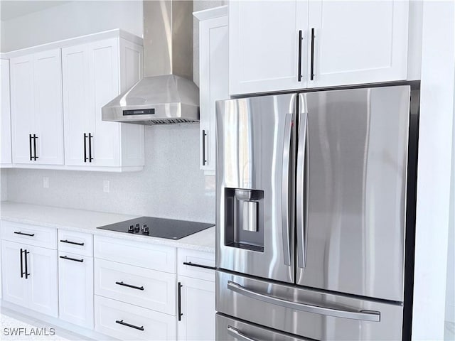 kitchen with light stone counters, wall chimney range hood, stainless steel fridge, black electric stovetop, and white cabinets