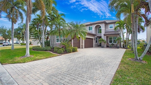 mediterranean / spanish-style home with a tile roof, stucco siding, an attached garage, a front lawn, and decorative driveway