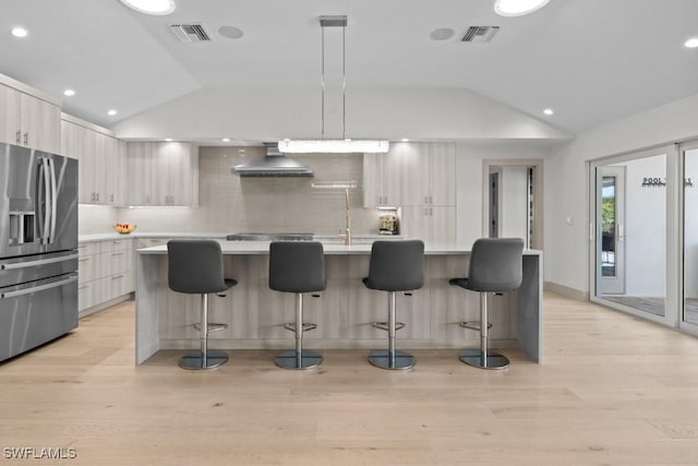 kitchen with an island with sink, lofted ceiling, stainless steel fridge, hanging light fixtures, and wall chimney range hood