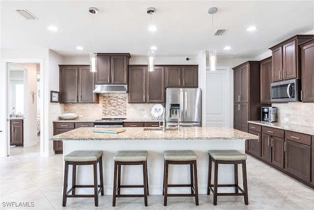 kitchen featuring appliances with stainless steel finishes, decorative light fixtures, an island with sink, light stone countertops, and dark brown cabinets