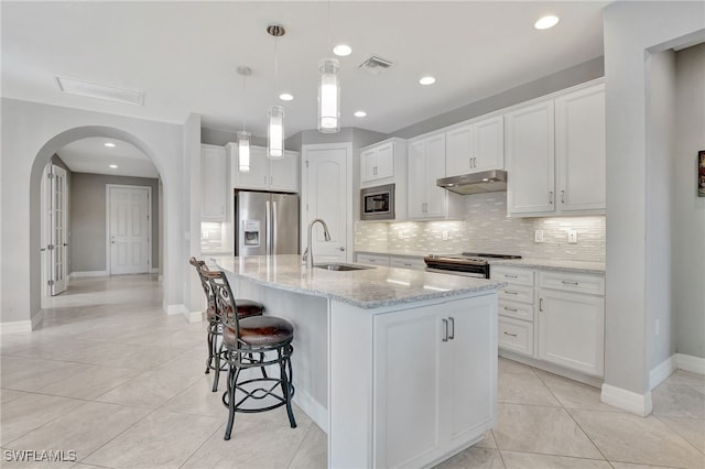 kitchen with appliances with stainless steel finishes, decorative light fixtures, white cabinets, light stone counters, and a center island with sink