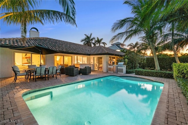 pool at dusk with an outdoor pool, a patio, an outdoor kitchen, and an outdoor hangout area