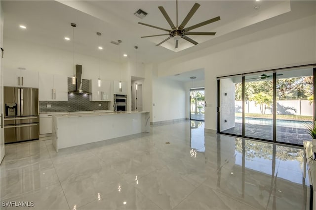 kitchen featuring appliances with stainless steel finishes, white cabinetry, an island with sink, hanging light fixtures, and wall chimney exhaust hood