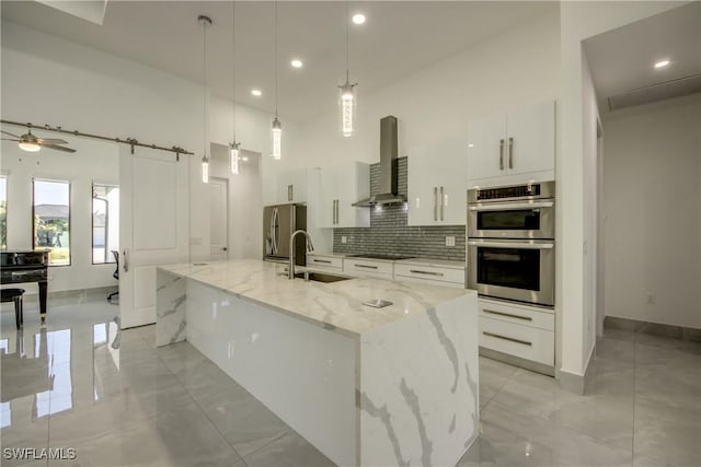 kitchen with wall chimney range hood, sink, a kitchen island with sink, hanging light fixtures, and white cabinetry