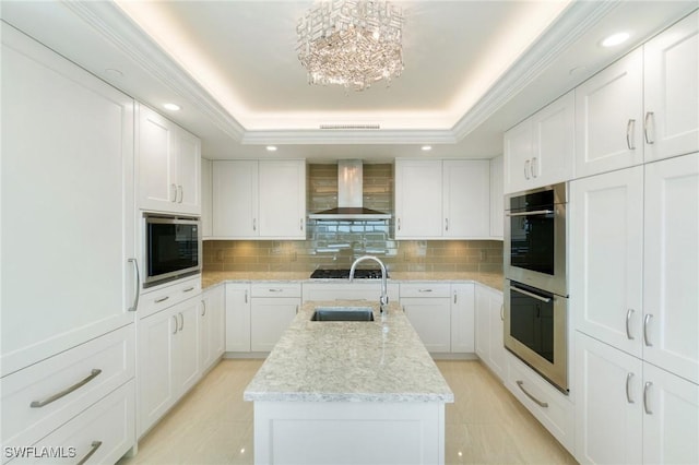 kitchen with a raised ceiling, wall chimney exhaust hood, a kitchen island with sink, built in microwave, and double oven