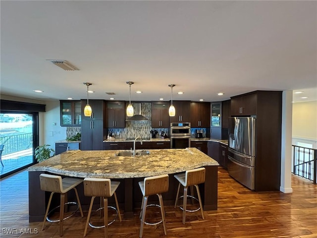 kitchen with wall chimney range hood, hanging light fixtures, dark brown cabinets, stainless steel appliances, and an island with sink