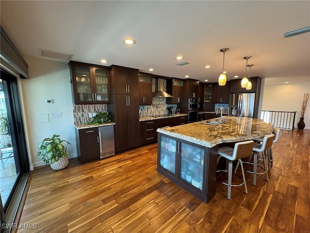 kitchen with wall chimney exhaust hood, sink, dark brown cabinets, an island with sink, and stainless steel appliances