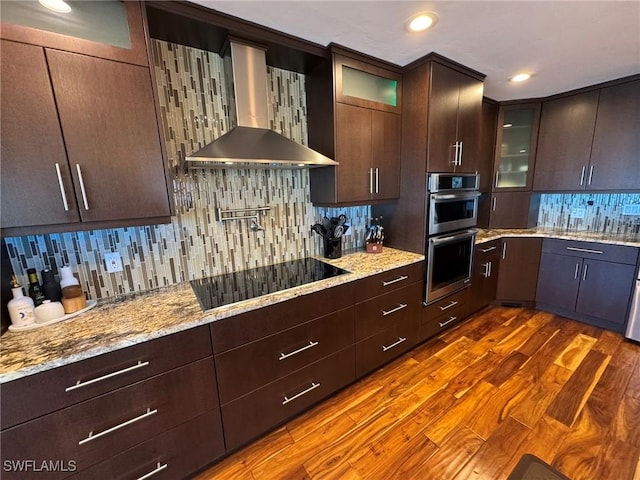 kitchen with black electric stovetop, stainless steel double oven, wall chimney exhaust hood, and light stone countertops