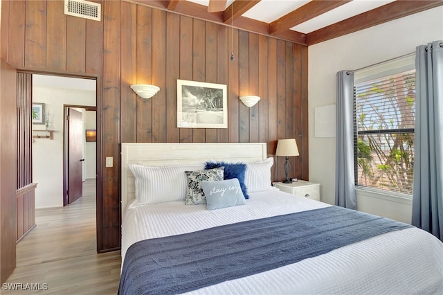 bedroom with multiple windows, beam ceiling, light hardwood / wood-style flooring, and wood walls