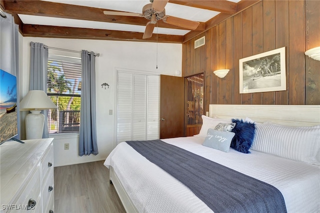 bedroom with ceiling fan, light wood-type flooring, a closet, beamed ceiling, and wood walls