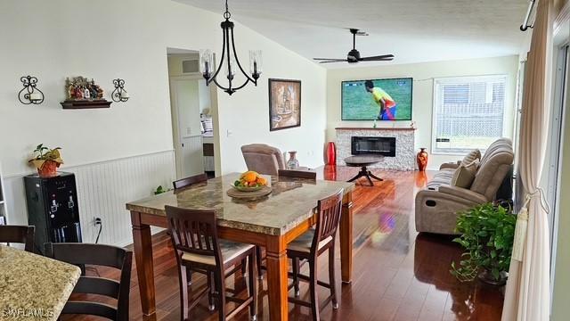 dining area featuring wainscoting, a glass covered fireplace, wood finished floors, vaulted ceiling, and ceiling fan with notable chandelier