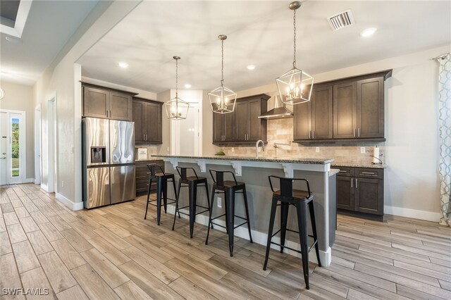 kitchen featuring a kitchen island with sink, dark brown cabinetry, stainless steel refrigerator with ice dispenser, light stone countertops, and wall chimney exhaust hood