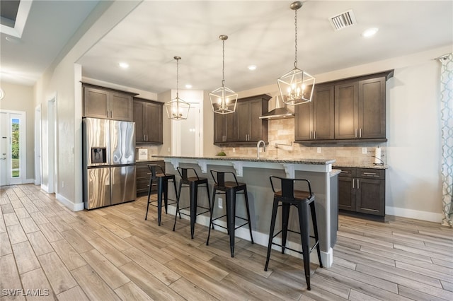 kitchen with wall chimney range hood, stainless steel fridge, dark brown cabinetry, light stone counters, and a center island with sink