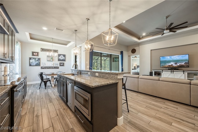 kitchen with appliances with stainless steel finishes, a tray ceiling, light stone countertops, a center island with sink, and decorative light fixtures