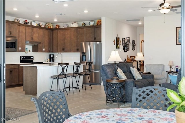 kitchen featuring a breakfast bar area, dark brown cabinets, an island with sink, ceiling fan, and stainless steel appliances