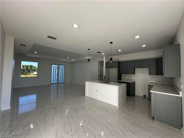 kitchen featuring sink, gray cabinetry, decorative light fixtures, a raised ceiling, and a kitchen island with sink