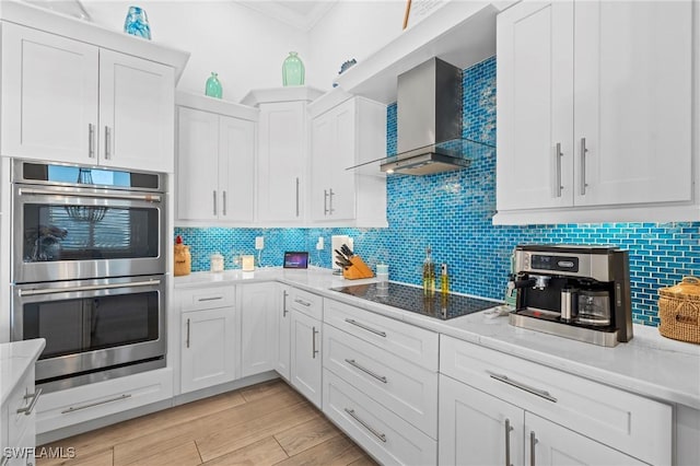 kitchen with wall chimney exhaust hood, white cabinetry, stainless steel double oven, black electric stovetop, and light stone countertops