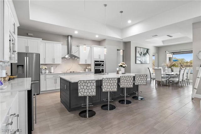 kitchen featuring stainless steel appliances, decorative backsplash, a kitchen bar, a raised ceiling, and wall chimney range hood