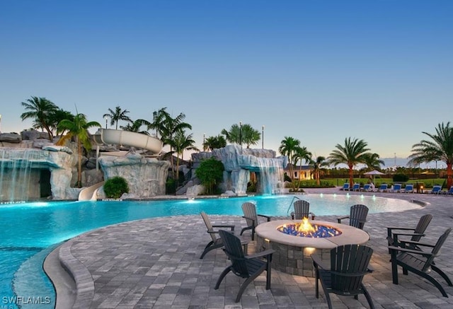 pool at dusk featuring a patio area, a water slide, a fire pit, and pool water feature