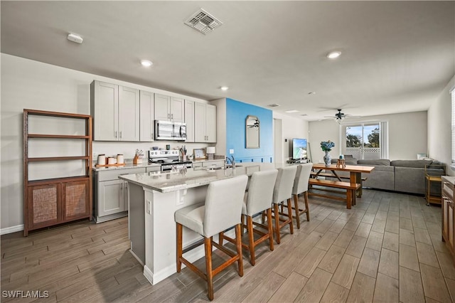 kitchen with a breakfast bar, an island with sink, ceiling fan, stainless steel appliances, and light stone countertops