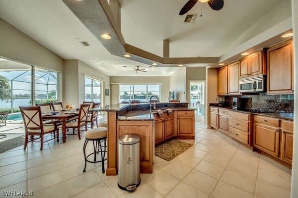 kitchen featuring a breakfast bar area, a tray ceiling, an island with sink, ceiling fan, and dark stone counters