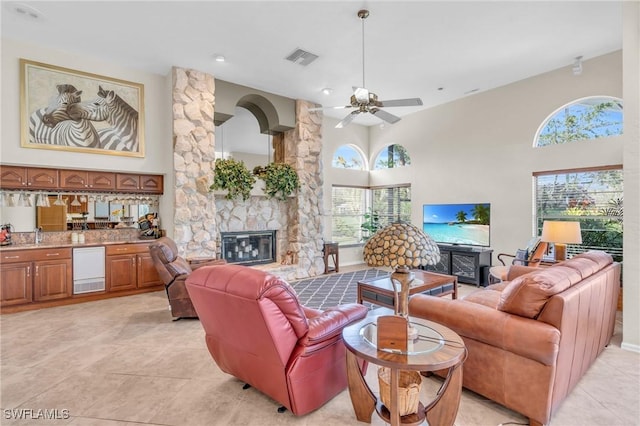 tiled living room with ceiling fan, a healthy amount of sunlight, a stone fireplace, and a high ceiling