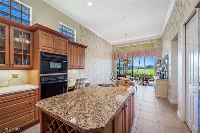 kitchen with decorative light fixtures, an island with sink, sink, ornamental molding, and light stone counters
