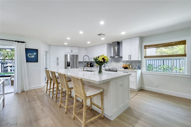 kitchen featuring white cabinetry, an island with sink, sink, stainless steel refrigerator with ice dispenser, and wall chimney range hood