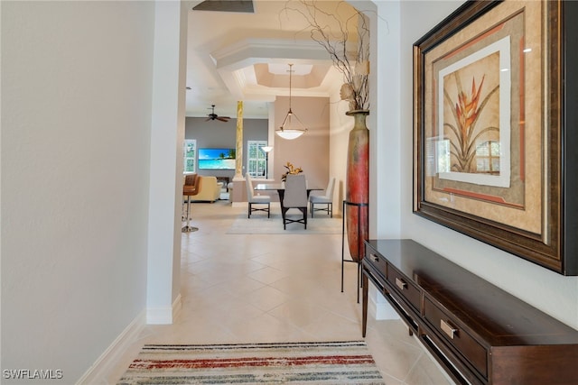 hallway featuring a tray ceiling, baseboards, light tile patterned floors, and ornamental molding