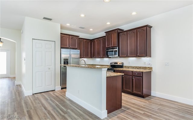 kitchen featuring an island with sink, dark brown cabinetry, light stone counters, stainless steel appliances, and light hardwood / wood-style floors