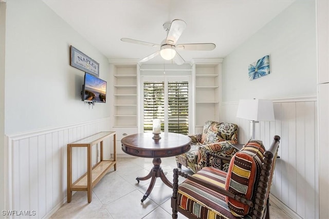 living area with built in shelves, a wainscoted wall, ceiling fan, and light tile patterned floors