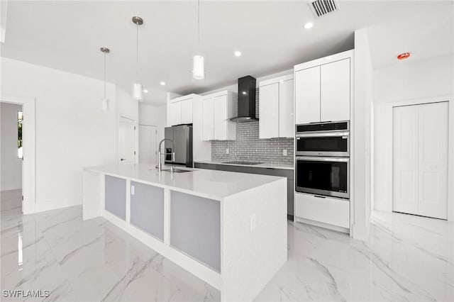 kitchen featuring wall chimney range hood, an island with sink, pendant lighting, stainless steel appliances, and white cabinets