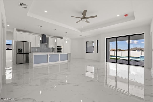 kitchen featuring pendant lighting, white cabinets, a tray ceiling, stainless steel appliances, and wall chimney exhaust hood