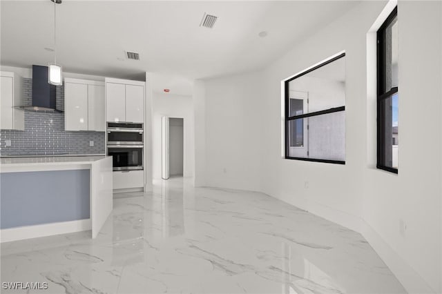 kitchen with tasteful backsplash, white cabinets, pendant lighting, stainless steel double oven, and wall chimney range hood