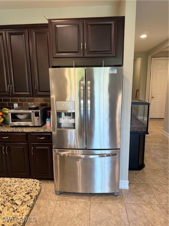 kitchen with tasteful backsplash, dark brown cabinets, light tile patterned floors, stainless steel fridge, and light stone countertops