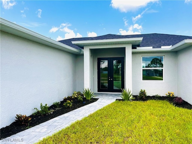 view of exterior entry with french doors, roof with shingles, a lawn, and stucco siding