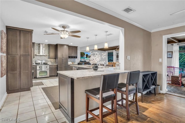 kitchen with hanging light fixtures, dark brown cabinetry, stainless steel electric range oven, wall chimney range hood, and a breakfast bar area