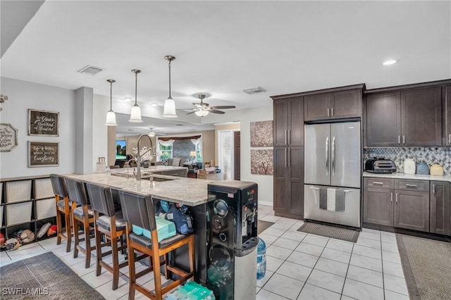 kitchen featuring a center island with sink, light tile patterned flooring, stainless steel fridge, hanging light fixtures, and decorative backsplash