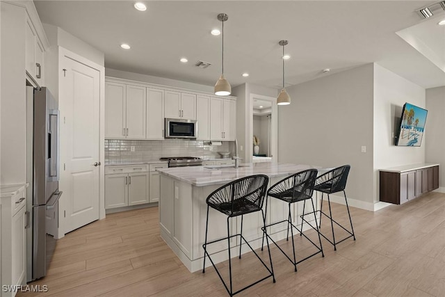 kitchen with white cabinetry, hanging light fixtures, an island with sink, stainless steel appliances, and light hardwood / wood-style floors