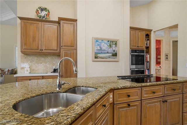 kitchen with double oven, light stone countertops, a sink, decorative backsplash, and brown cabinetry