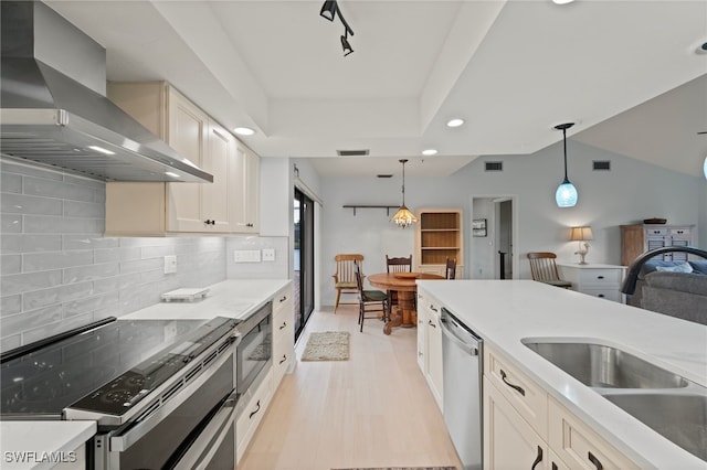 kitchen with hanging light fixtures, stainless steel appliances, sink, a tray ceiling, and wall chimney exhaust hood