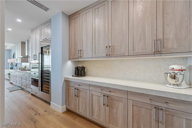 kitchen featuring wall chimney range hood, backsplash, light hardwood / wood-style flooring, and light brown cabinets