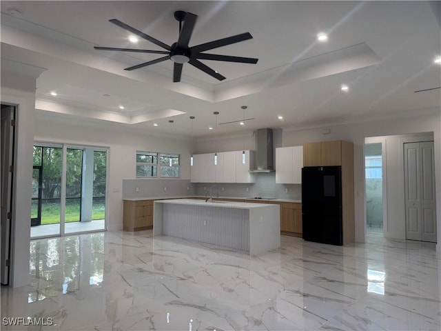 kitchen featuring white cabinetry, a center island with sink, a tray ceiling, wall chimney range hood, and black appliances