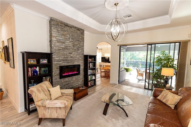 living room featuring a stone fireplace, ornamental molding, a raised ceiling, a notable chandelier, and light hardwood / wood-style floors